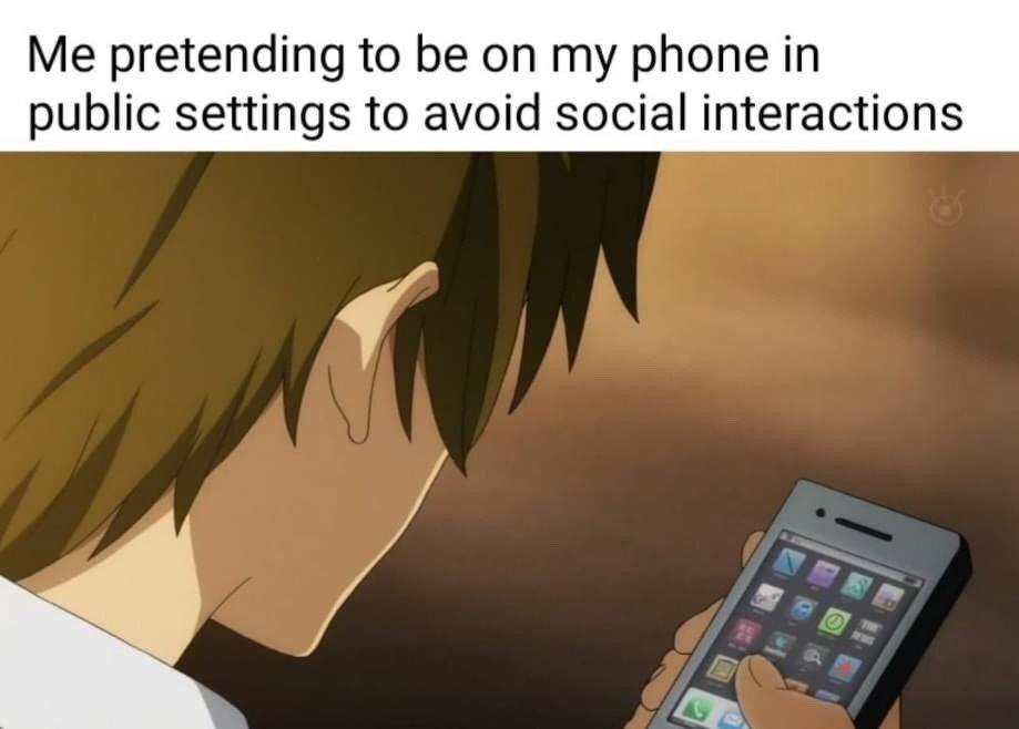 This meme consists of top overlaid text paired with an anime screenshot below. The text reads 'Me pretending to be on my phone in public settings to avoid social interactions'. The visual shows the back of a young male anime character holding a smartphone and staring intently at its screen, appearing occupied with the device. It illustrates the common relatable behavior of faking phone usage to avoid unwanted small talk or interactions with other people when in public spaces.