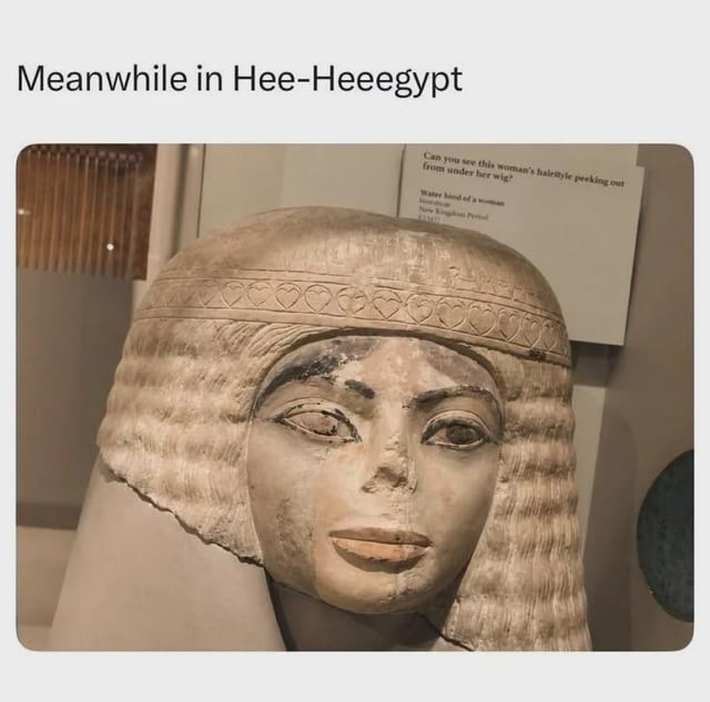 This is a single-panel photo meme. The main image is a close-up shot of an ancient Egyptian carved limestone female bust from the New Kingdom period, on display in a museum. The bust wears a layered wavy wig topped with a carved headband decorated with repeating heart motifs, and its painted facial features have a faint subtle smile. An information plaque for the exhibit is visible behind the statue. Above the photo, a caption reads "Meanwhile in Hee-Heeegypt". The core joke is a pun combining pop singer Michael Jackson's iconic signature "Hee-Hee" vocal ad-lib with the word "Egypt", as the statue's facial expression is humorously compared to the face Jackson makes when performing the ad-lib, implying this ancient artifact is an early predecessor of Jackson's famous mannerism.