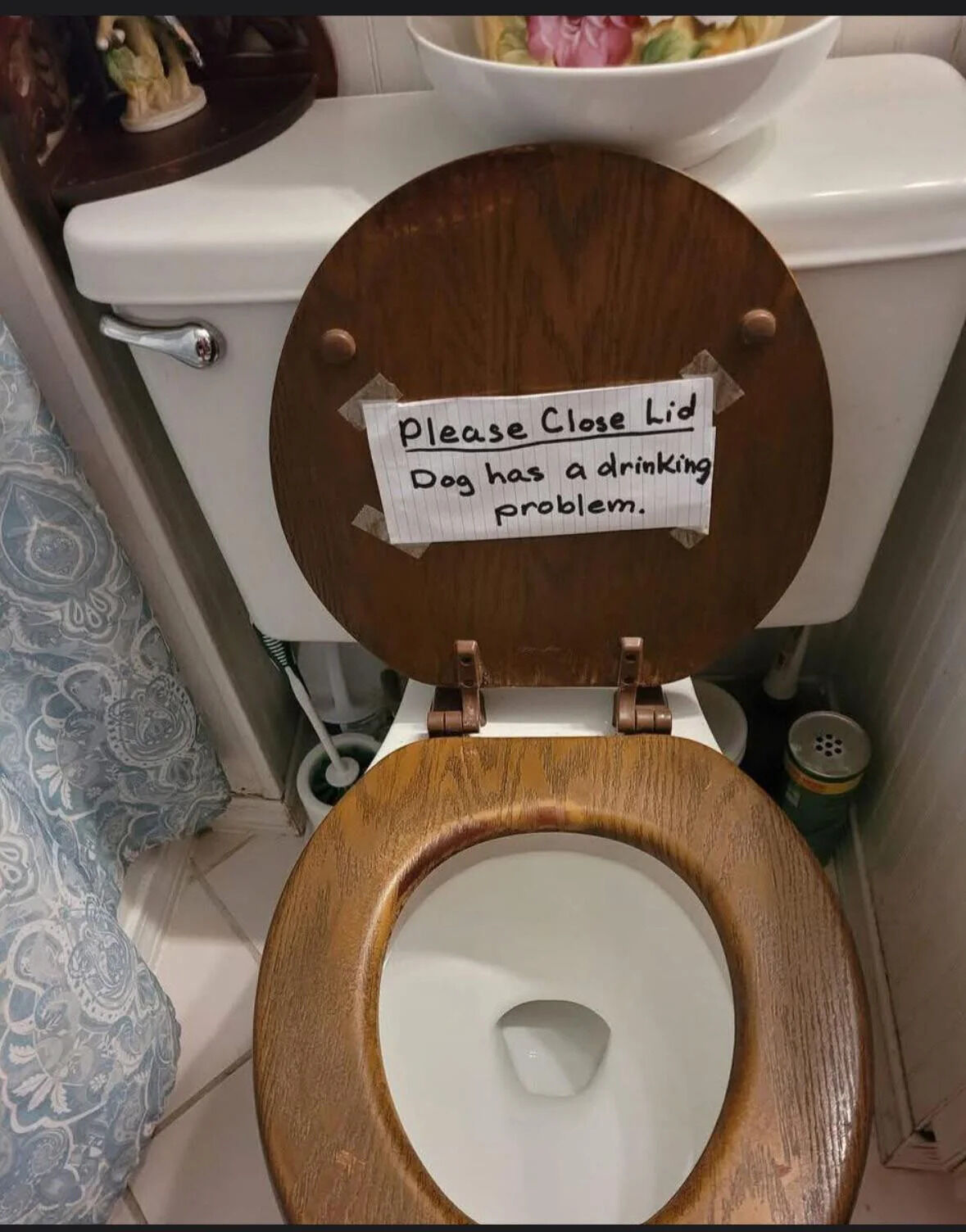 A photograph of a bathroom toilet with a wooden lid propped open. Affixed to the lid with tape is a handwritten note on lined paper that reads: 'Please Close Lid Dog has a drinking problem.' The toilet is white with a chrome flush handle. Visible in the surrounding bathroom are a floral-patterned shower curtain to the left, a toilet brush with a white holder beneath the toilet, and a decorative bowl with artificial flowers placed on top of the toilet tank. The scene is a typical residential bathroom, capturing a humorous household solution to a pet-related issue.