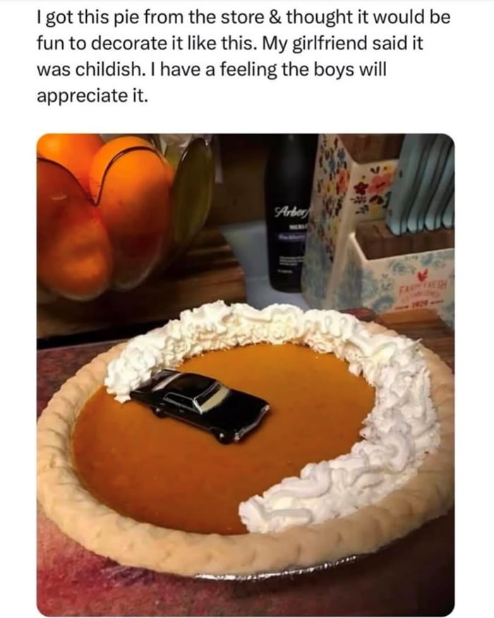 A meme consisting of a text overlay and a photograph. The text reads: 'I got this pie from the store & thought it would be fun to decorate it like this. My girlfriend said it was childish. I have a feeling the boys will appreciate it.' Below the text is a color photograph of a pumpkin pie with an orange filling, surrounded by a border of white whipped cream. In the center of the pie sits a small black toy car. The background features kitchen items including a wooden cutting board with a bowl of oranges, a bottle labeled 'Arbor Mist', and a floral-patterned utensil holder with the text 'Farm Fresh' visible.