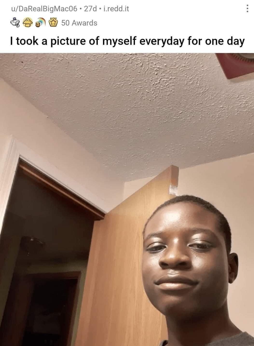 This is a screenshot of a viral Reddit post. The post's caption reads 'I took a picture of myself everyday for one day', an absurdist technically correct joke that subverts the common trend of people taking daily self-portraits over extended periods of weeks, months, or years. Below the caption is a low-angle selfie of a young Black man with a calm, slightly amused expression, standing in a residential indoor setting with a wooden door, textured popcorn ceiling, and a dark doorway leading to another room visible in the background. The original Reddit post received 50 awards from other users.