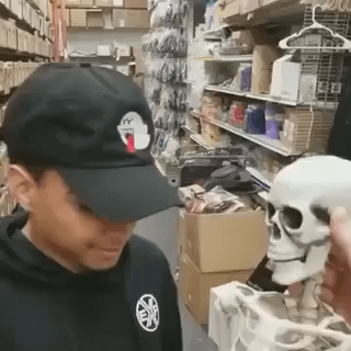 This is a casual photograph taken inside a cluttered storage space or secondhand thrift store. On the left, a man wearing a black baseball cap and black hoodie stands looking downward. On the right, an unseen person's hand holds up a full-size articulated plastic human skeleton model, which is an unusual unexpected find. The original post asks viewers to guess what the strange find is, with the skeleton being the surprising item discovered here.
