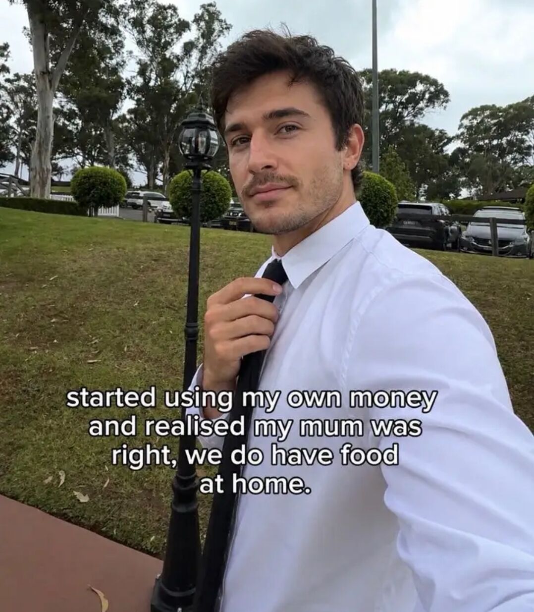 The image features a young adult man wearing a white dress shirt and black tie, standing outdoors next to a decorative lamppost on a grassy area with trees and parked cars visible in the background. He is adjusting his tie while looking at the camera with a knowing, resigned expression. Overlaid on the lower part of the image is text that reads: "started using my own money and realised my mum was right, we do have food at home." The joke is relatable to most young adults, referencing the common childhood habit of asking for takeout or eating out, only to understand once they start paying for their own expenses that eating food already available at home is a cheaper, practical option that their mothers were correctly advising them to choose.