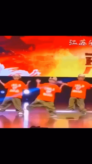 This still image captures a moment from a children's group dance performance on a professional stage. Three young boys wearing matching orange printed t-shirts, khaki pants, and light-colored caps are performing a synchronized dance move with their arms outstretched and legs spread wide. The core comedic element of the meme is that the middle boy's pants are visibly slipping down his hips, yet he continues performing the routine without pausing, which aligns with the title's reference to the common show business saying. The stage background features a bright red-to-yellow gradient graphic with white wave illustrations, partial Chinese characters are visible on the upper right of the backdrop, and the stage has a reflective floor with active stage lighting.