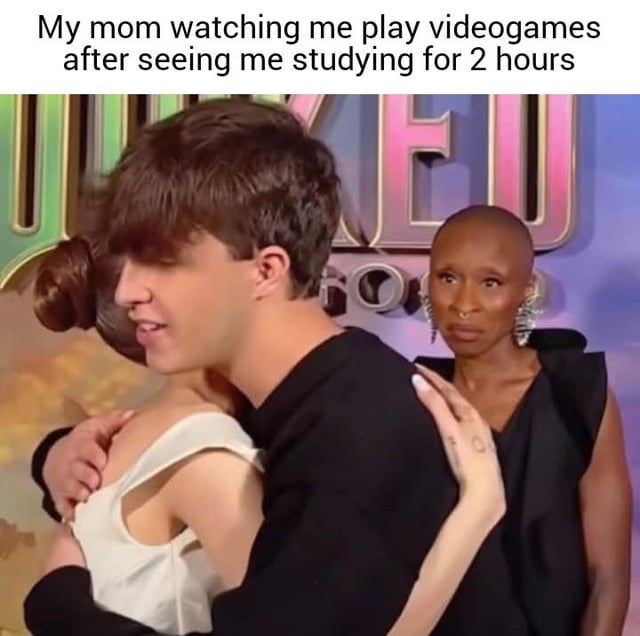 A meme image featuring three real people. In the foreground, a man in a black shirt hugs a woman in a white dress. In the background, another woman with a bald head, wearing a black outfit and large earrings, stands with a disapproving or judging expression. The top text reads: 'My mom watching me play videogames after seeing me studying for 2 hours', where the background woman represents the mom reacting to the 'me' (implied to be the hugging man or woman) playing games after studying.
