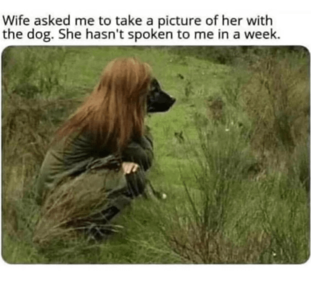 A meme image featuring a photograph of a woman with long red hair crouching in a grassy, overgrown field. She is holding a black dog close to her chest, with both facing away from the camera. Above the image, a text caption reads: 'Wife asked me to take a picture of her with the dog. She hasn't spoken to me in a week.' The humor stems from the implied narrative that the husband's failure to take a satisfactory photo (e.g., capturing them from behind instead of facing the camera) has led to his wife's prolonged silence, highlighting relatable relationship mishaps.