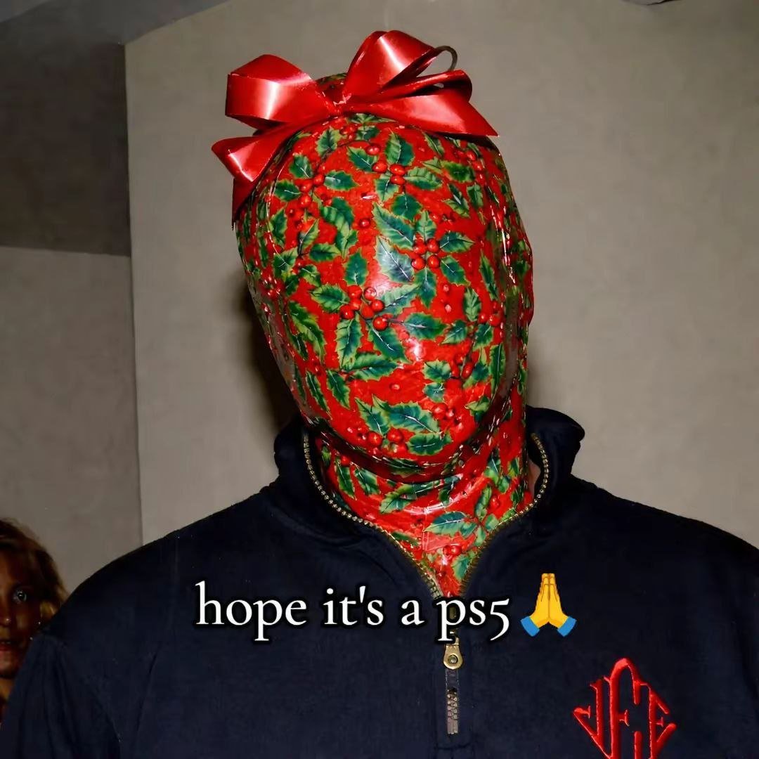 A person with their entire head covered in red wrapping paper adorned with green holly leaves and red berries, topped with a large red bow, resembling a Christmas gift. They are wearing a dark-colored zip-up jacket. Overlaid text at the bottom reads 'hope it's a ps5' accompanied by a praying hands emoji 🙏, humorously implying the wrapped head is a 'gift' hoping to contain a PS5 console.