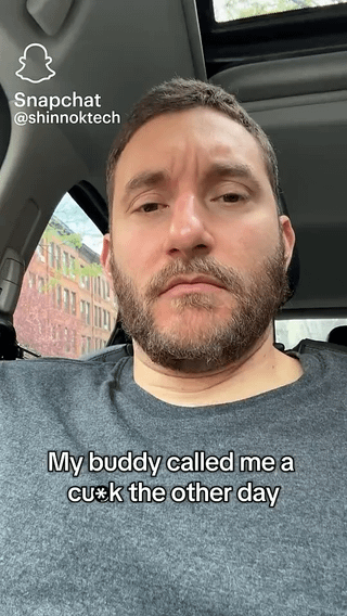 This is a first-person selfie screenshot of a bearded white man with short brown hair, sitting inside a car. He has a deadpan, unamused expression as he looks directly at the camera. Overlaid white text on the lower portion of the image reads "My buddy called me a cu*k the other day". A Snapchat watermark with the creator's username @shinnoktech is visible in the top left corner, and a brick building can be seen through the car window behind the man. This frame is the opening of a short video that teases a longer humorous story about the insult.