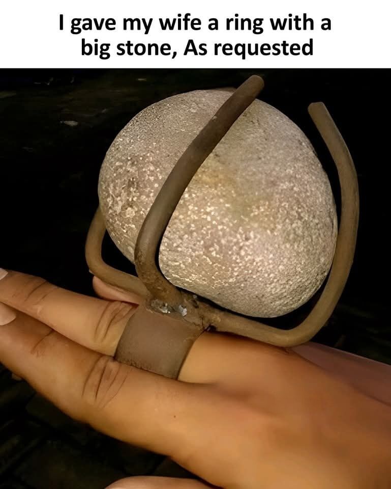 The image displays a person's outstretched hand wearing a roughly constructed oversized metal ring. Instead of holding a traditional precious gemstone, the ring is built to hold an entire palm-sized smooth natural river rock. Above the photograph, text states 'I gave my wife a ring with a big stone, As requested'. The core humor comes from a literal pun: when people ask for a ring with a 'big stone', they typically mean a large valuable gem like a diamond, but the creator of the joke took the request completely literally and used an actual large rock, resulting in an absurd, lighthearted humorous gift.