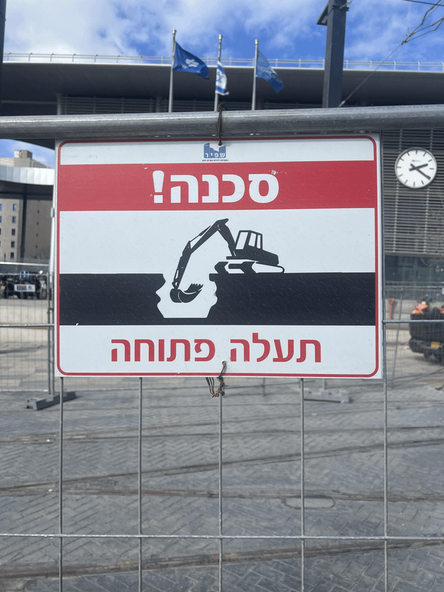 The image displays an official public safety warning sign mounted on a metal fence at an urban construction site. The sign uses Hebrew text: the top red bar reads 'סכנה!' (Danger!), the central section features a black-and-white graphic of an excavator positioned over an open pit, and the bottom bar reads 'תעלה פתוחה' (Open Excavation). A small watermark for 'שימור - חברת הכבישים הלאומית לישראל' (Shimur, National Roads Company of Israel) is visible at the top center of the sign. In the background, there is a modern multi-story building with three flags (two blue with white emblems, one white with a blue emblem), a clock showing roughly 10:10, and construction equipment, confirming an active worksite environment.