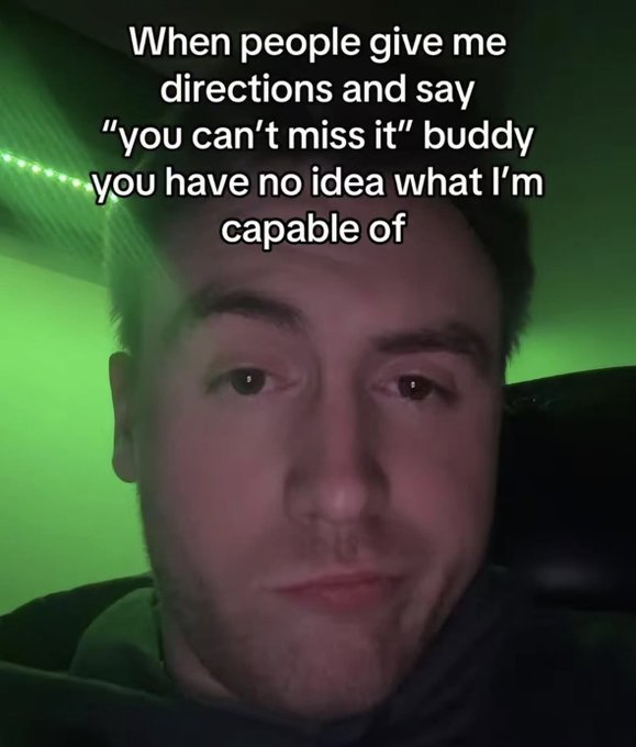 The image is a low-light selfie of a young man with a neutral, deadpan expression, lit by green ambient light from what appears to be the interior of a car. White text is overlaid across the top of the image that reads "When people give me directions and say 'you can't miss it' buddy you have no idea what I'm capable of". The joke is a self-deprecating punchline that plays on relatable experiences of being directionally challenged, implying the poster is so bad at navigation they absolutely will miss the referenced location even if it is extremely obvious and easy to find for most people.