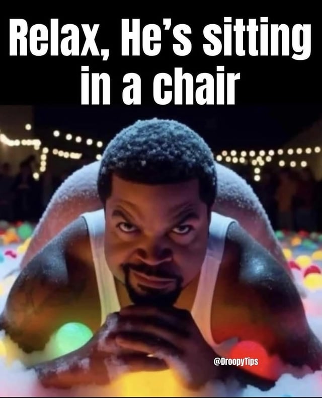 This meme displays bold white text on a black header that reads 'Relax, He's sitting in a chair'. Below the header is a low-angle shot of rapper and actor Ice Cube, who has a light frosty dusting on his hair and shoulders, wears a white tank top, and stares intensely at the viewer with an angry, menacing scowl, his hands clasped together in front of him. He appears to be positioned in a ball pit with colorful plastic balls, with out-of-focus string lights in the background. The forced perspective of the shot makes Ice Cube look like he is looming aggressively very close to the viewer, creating a comedic contrast with the caption that clarifies he is simply performing the ordinary, non-threatening action of sitting in a chair. A watermark reading '@DroopyTips' is visible in the bottom right corner of the image.