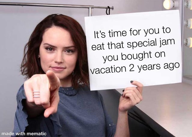 A meme image featuring a woman with shoulder-length brown hair wearing a blue top. She is pointing directly at the viewer with her right hand while holding a white sign in her left hand. The sign displays the text: 'It's time for you to eat that special jam you bought on vacation 2 years ago'. The background shows a vanity area with a mirror and light bulbs. A 'made with mematic' watermark is visible at the bottom of the image. The scene humorously addresses the relatable experience of holding onto unused special food items purchased during travel.