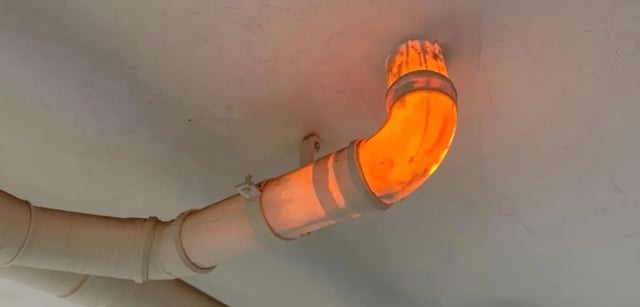 The image displays a section of white plumbing pipe mounted on a grey ceiling, with a curved portion of the pipe glowing bright fiery orange as if it is extremely hot or contains burning material. The meme uses this visual to make an exaggerated, humorous joke: it implies that the upstairs neighbor ate extremely spicy Korean Buldak (fire) noodles, and the resulting waste passing through the shared plumbing is so hot and spicy that it made the pipe glow orange, leaning into the well-known reputation of Buldak noodles for being exceptionally spicy.