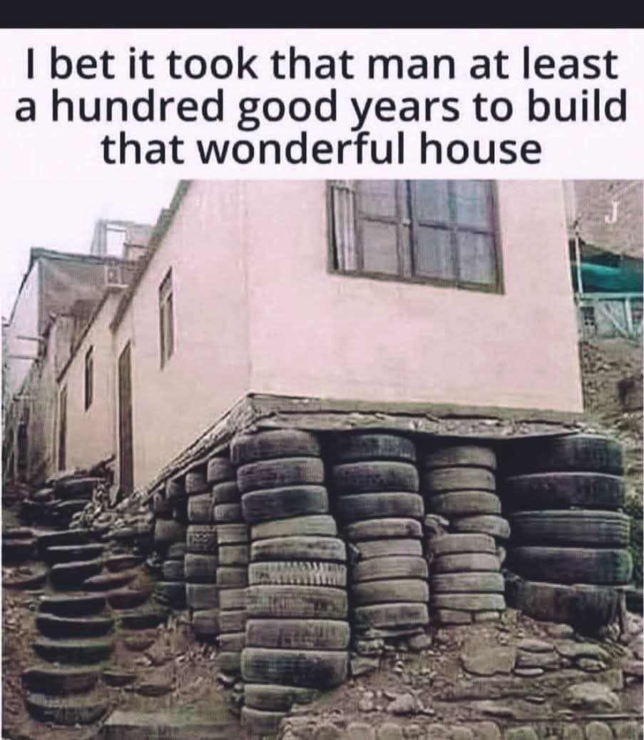 A photograph of a house constructed on a sloped terrain, featuring a unique foundation made from stacked used tires. The house has a light pink exterior with multiple rectangular windows and appears to be part of a hillside settlement. Above the house, a text overlay reads, 'I bet it took that man at least a hundred good years to build that wonderful house,' which playfully exaggerates the time and effort inferred to have been invested in creating the resourceful and visually distinctive structure.