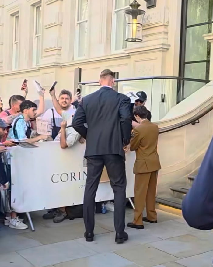 The image captures a scene outside the Corinthia London hotel, where a crowd of fans waits behind a barrier holding items to be signed and phones to take photos. Two men stand with their backs to the camera facing the crowd: one is an extremely tall, broad-shouldered man wearing a dark suit with short cropped hair, the other is a shorter man in a tan suit. The meme asks viewers to spot the legendary Greek warrior: the answer is the tall man in the dark suit, whose silhouette from the back is nearly identical to that of Leonidas, the iconic Spartan Greek warrior from the film *300*.