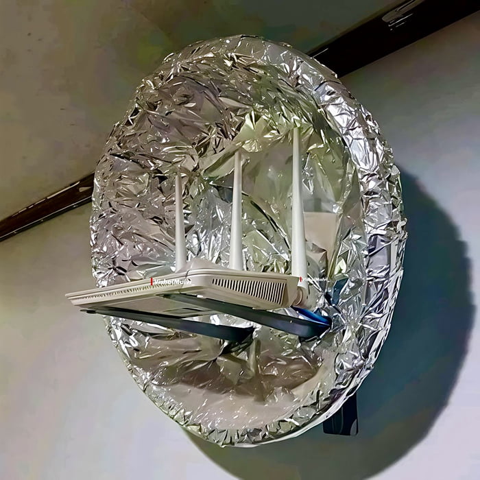 The image depicts a DIY Wi-Fi setup featuring a white Wi-Fi router with three vertical antennas mounted inside a large, crumpled aluminum foil dish. The foil dish, which appears to be roughly circular and attached to a wall, is likely intended as a homemade signal booster. The router is positioned such that its antennas extend outward from the foil dish, and the foil itself is wrinkled, reflecting light. The setup is mounted on a plain wall, with some structural elements (like a wooden beam) visible in the background.