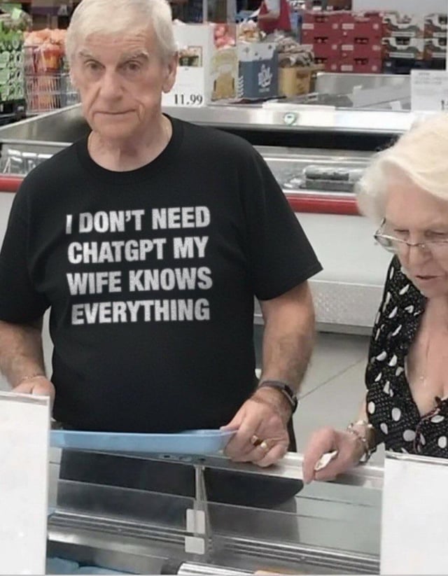 The image captures an elderly man at a supermarket checkout counter, wearing a black t-shirt printed with bold white text: 'I DON'T NEED CHATGPT MY WIFE KNOWS EVERYTHING'. Standing beside him is an elderly woman, presumably his wife, who is focused on the checkout area, seemingly unaware of the playful joke on the man's shirt. The background shows typical grocery store elements, including checkout equipment and product shelves. The humor stems from a relatable, lighthearted marital stereotype: framing the wife as an all-knowing source of information that renders the AI chatbot ChatGPT unnecessary. This taps into shared, playful jokes about spouses who claim to have all the answers, making the meme relatable to many people in long-term relationships.