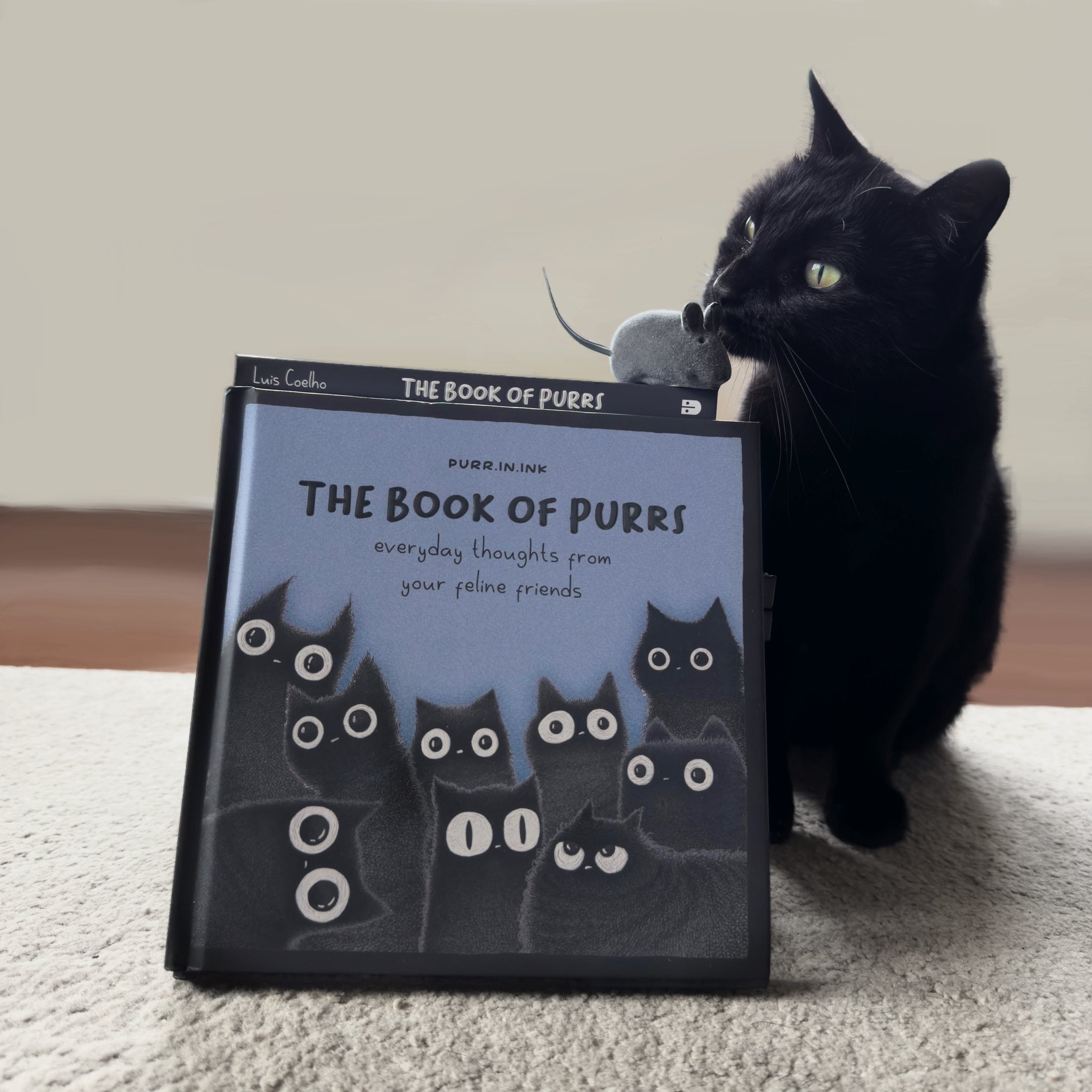 A black cat with striking green eyes sits on a light-colored carpet, holding a small gray toy mouse in its mouth. To the cat's left is a hardcover book titled 'THE BOOK OF PURRS' with a blue cover. The book's cover features the text 'PURR.INK' at the top, the main title 'THE BOOK OF PURRS' in bold black letters, and the subtitle 'everyday thoughts from your feline friends' below. The cover art consists of multiple illustrations of black cats with large, round eyes. The background is a plain, light-colored wall with a wooden floor visible at the bottom edge. The cat appears calm and engaged, with its head turned slightly toward the book.