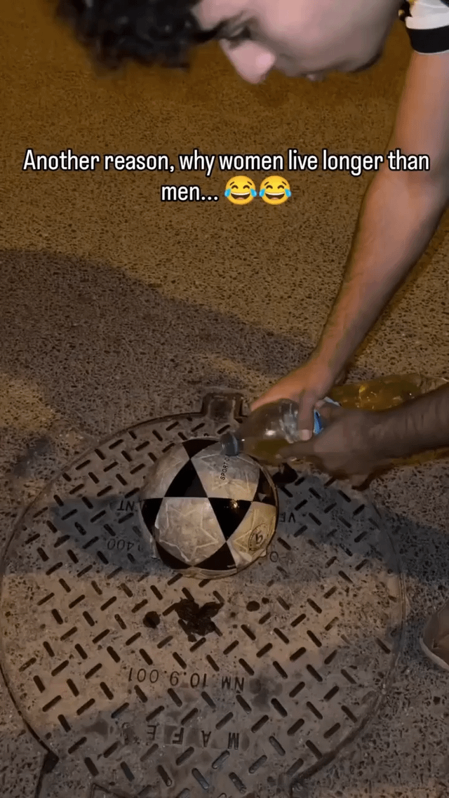 This meme features a real man leaning over a metal manhole cover, pouring a liquid (likely alcohol) onto a soccer ball resting on the manhole. The top text reads *"Another reason, why women live longer than men.... 😂😂"*, satirizing the common stereotype that men engage in more reckless, unnecessary risky behaviors that could lead to harm or shorten their lifespan. The humor comes from the absurdity of the action—there's no logical reason to pour liquid on a soccer ball on a manhole, framing it as an example of the dumb, risky choices men make.