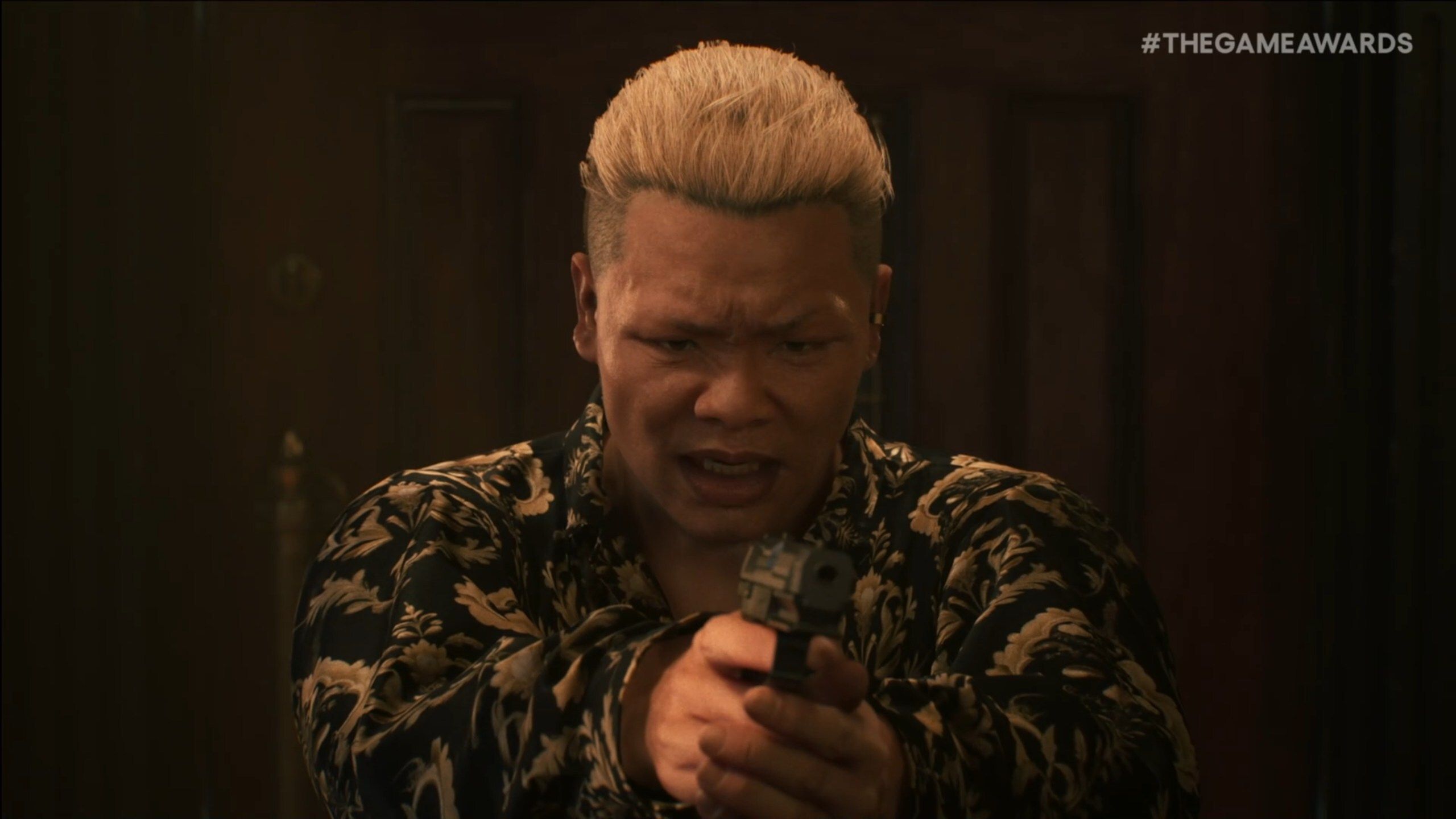 A screenshot featuring a real person with short blonde hair wearing a black and gold floral-patterned shirt. The individual is in a dimly lit room with wooden walls, holding a handgun pointed directly at the camera with both hands. Their expression is intense and angry, with furrowed brows and an open mouth, suggesting a dramatic or confrontational moment. A watermark '#THEGAMEAWARDS' is visible in the top right corner of the image.
