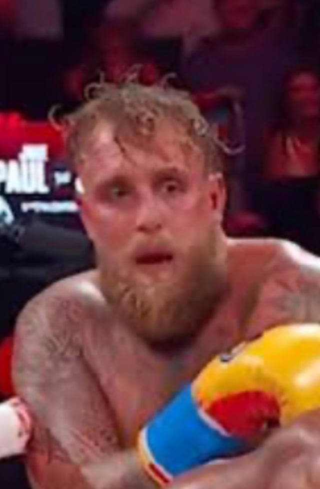 A close-up screenshot of a shirtless male boxer with a full beard, visible tattoos on his arms, and wearing yellow boxing gloves. The boxer appears sweaty with disheveled hair, displaying a shocked or surprised expression. He is positioned in a boxing ring, with a blurred audience and a partially visible 'PAUL' banner in the background, suggesting the event may be associated with Jake Paul or his promotion.