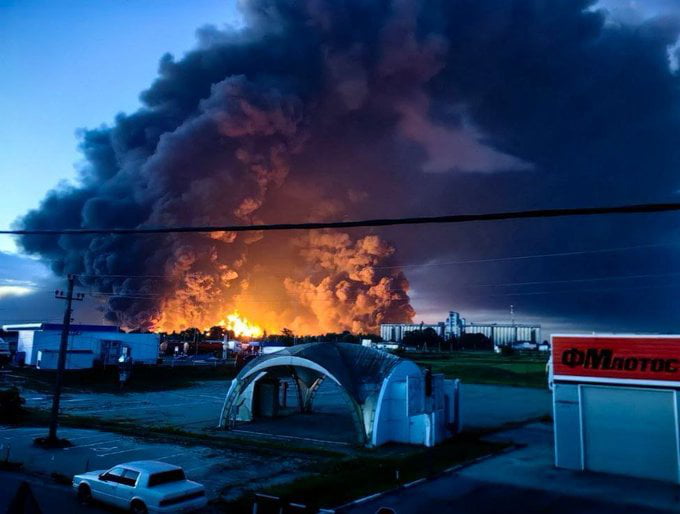 This image is a real photograph taken at dusk, depicting an enormous, blazing industrial fire in the distance, accompanied by huge billowing plumes of dark smoke lit orange from the flames. The foreground shows a low-density industrial/commercial area, including a parked white sedan, a semi-cylindrical arched portable structure, low-rise buildings, power lines, and a partially visible sign with Cyrillic text, confirming the location is in Russia. The accompanying post caption uses sarcastic dark humor, referring to the destructive fire, which is purported to be the result of Ukrainian strikes on two Russian oil refineries overnight, as 'beautiful scenery'.