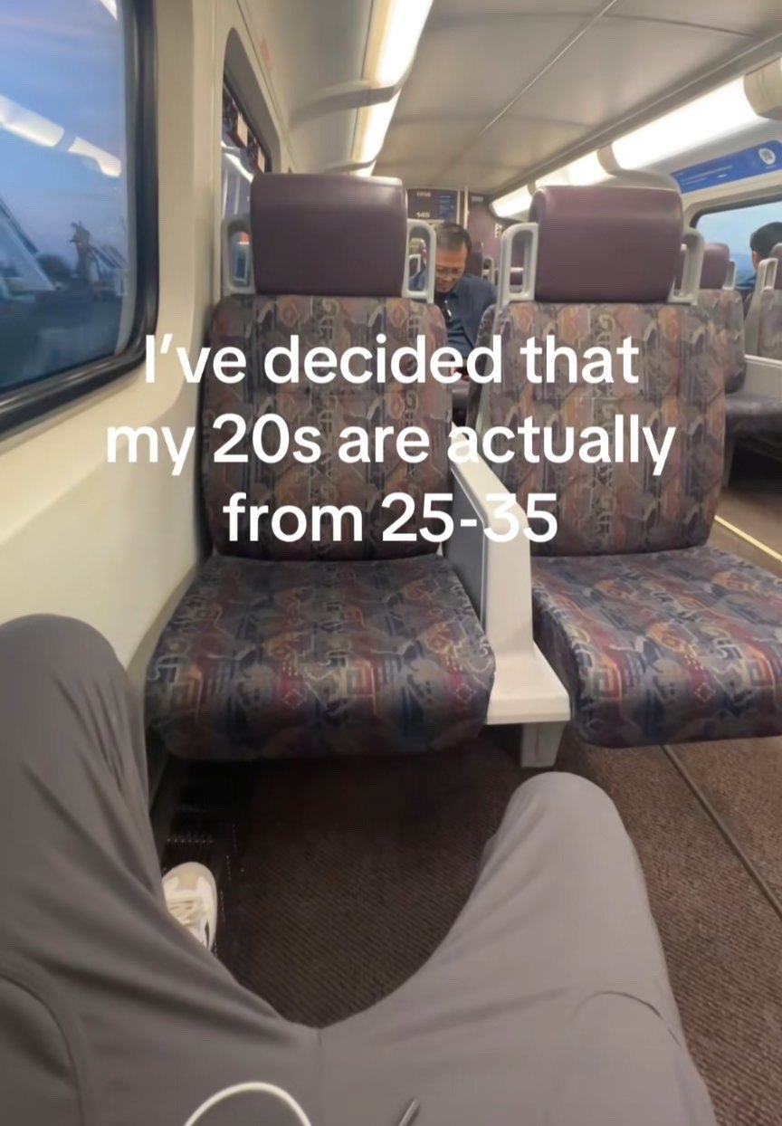 This is a first-person perspective photo taken inside a commuter train or rail car. The photographer's legs, clad in gray sweatpants with one white sneaker visible, are in the foreground. Two empty patterned upholstered train seats sit directly in front of the photographer, with other passengers visible further down the train carriage. Overlaid in white text across the center of the image is the phrase "I've decided that my 20s are actually from 25-35", which expresses a common relatable sentiment for modern young adults: rejecting traditional societal pressure to hit major life milestones by age 30, and redefining the period of young adulthood to account for economic and social barriers that delay typical milestones for many people today.