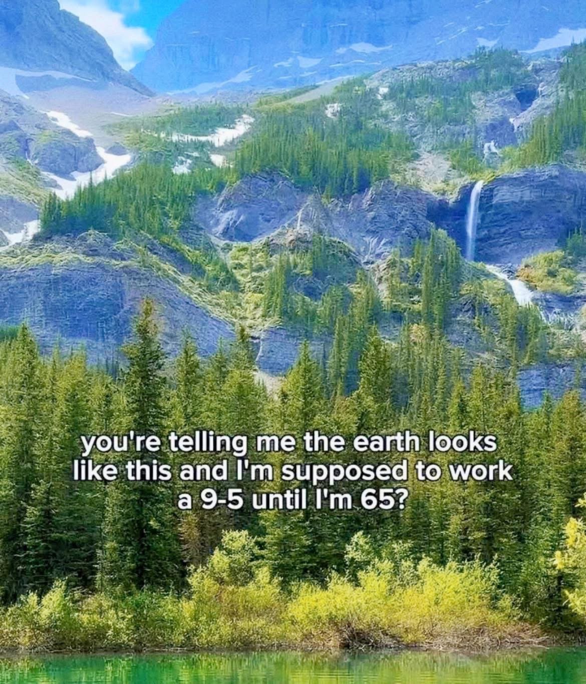 The meme features a breathtaking, vivid natural landscape as its background: towering, snow-dusted rocky mountain ranges rise in the distance, with a cascading waterfall running down one of the cliff faces, sloping hills covered in dense dark green coniferous forests, and a clear, bright turquoise-green lake in the immediate foreground. Overlaid in bold white sans-serif text across the forested middle section of the image is a rhetorical question expressing frustration with modern work norms: 'you're telling me the earth looks like this and I'm supposed to work a 9-5 until I'm 65?'. The joke relies on the contrast between the stunning, freeing beauty of the natural world and the restrictive, repetitive nature of traditional full-time employment that most people are expected to adhere to for most of their adult lives.