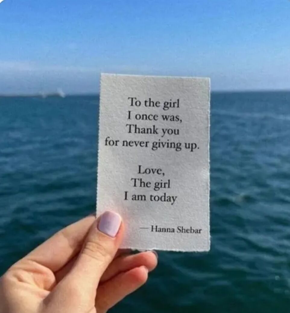 A photograph showing a hand with light purple nail polish holding a piece of white paper against an ocean backdrop. The paper contains a heartfelt message: 'To the girl I once was, Thank you for never giving up. Love, The girl I am today — Hanna Shebar'. The background features calm blue water and a clear sky with distant horizon.