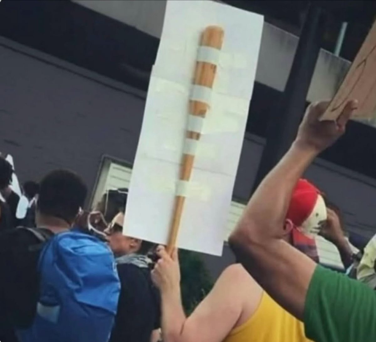The image captures a scene at a public gathering, likely a protest, where an attendee holds up a homemade sign consisting of a blank white rectangular sheet of paper attached to a wooden dowel with multiple evenly spaced pieces of white tape running along the length of the dowel. The entire setup creates an extremely convincing optical illusion that the real-world scene is a digital web page, as the sign and dowel perfectly mimic the appearance of a vertical web page scroll bar. Other protest attendees are visible in the surrounding area, including a person wearing a blue backpack facing away from the camera, a person in a red and white baseball cap holding up a different sign, and additional bystanders near a grey industrial-style building in the background. The humor of the image comes from the unexpected overlap between familiar digital user interface elements and real-world protest signage.