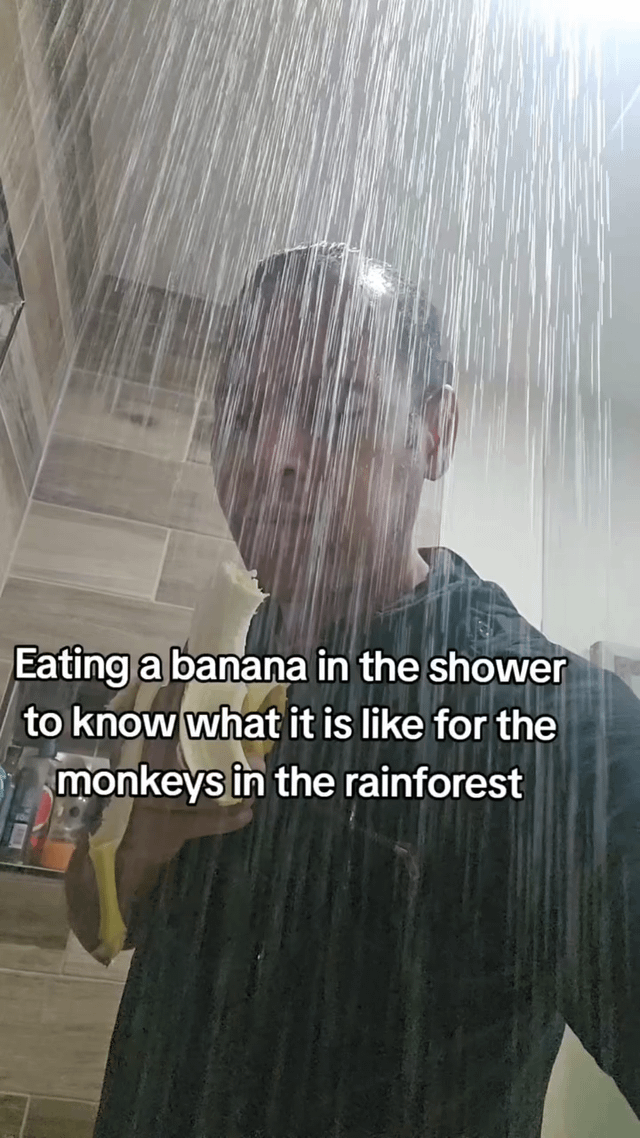 A humorous image showing a person standing in a shower with water pouring down, holding and eating a banana. The person appears to be male with short dark hair, wearing a dark-colored shirt. The shower has tiled walls and various toiletries are visible on a shelf in the background. Overlaid text on the image reads: 'Eating a banana in the shower to know what it is like for the monkeys in the rainforest'. The humor comes from the absurd premise of trying to experience what rainforest monkeys feel like by eating a banana in a shower (simulating rain).