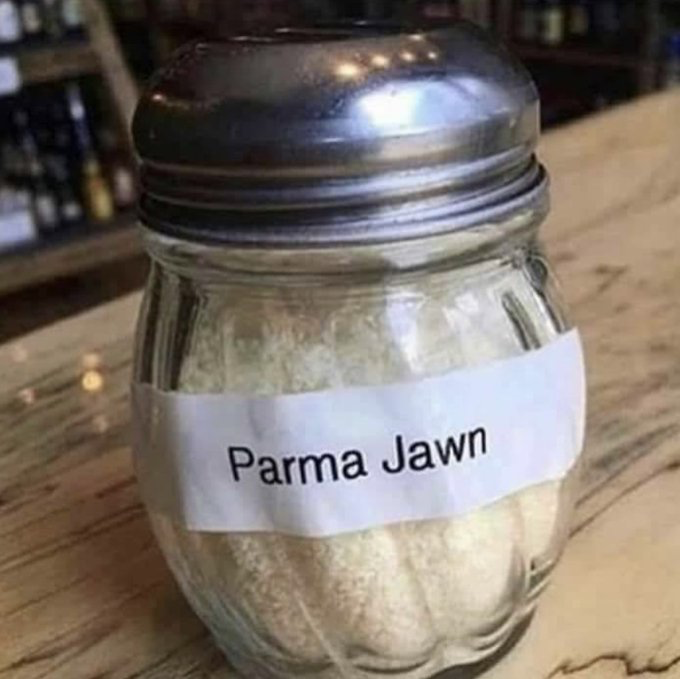 The image displays a standard glass grated parmesan cheese shaker with a perforated metal lid, placed on a light wooden restaurant counter. The shaker is filled with pale grated parmesan cheese, and has a plain white printed label affixed to it that reads "Parma Jawn". The humor comes from a phonetic pun: "Parma Jawn" sounds nearly identical to "Parmesan", and "jawn" is a widely used regional slang term originating from Philadelphia, USA that is used to refer to any unspecified object, place, or person, making the label a silly, regionally specific rebrand of the common condiment shaker.