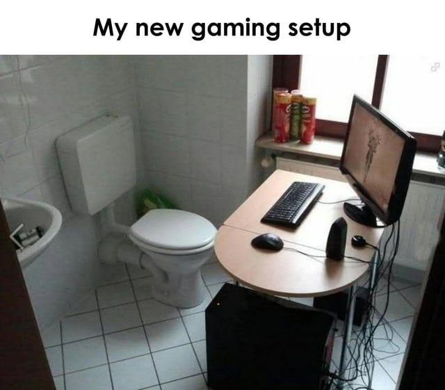 The image depicts a bathroom repurposed as a gaming area. Visible elements include a white toilet with a green object beside it, a small round desk holding a computer setup (monitor, keyboard, mouse, and speakers), a sink with toiletries, and several soda cans on a windowsill. The bathroom has white tiled walls and floor, creating a utilitarian setting. The humor stems from the unexpected and unconventional location (a bathroom) for a gaming setup, contrasting with typical elaborate or dedicated gaming spaces.