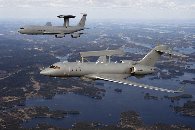 The image is an aerial photograph showing two Airborne Warning and Control System (AWACS) aircraft flying in formation over a landscape dotted with lakes and forested islands. The smaller aircraft in the foreground is the Swedish Saab GlobalEye, while the larger aircraft in the background marked 'U.S. AIR FORCE' is the American E-3 Sentry AWACS. The image is paired with the news that NATO has selected the Saab GlobalEye to replace its current fleet of 14 E-3 AWACS planes, representing a shift away from US-manufactured aircraft for this capability.