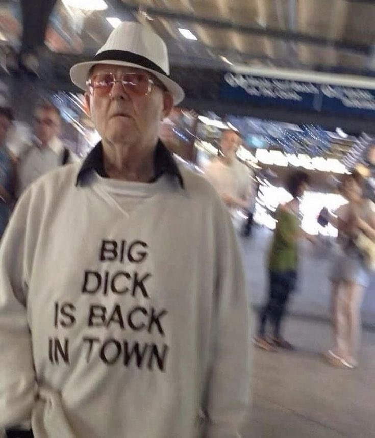 The image features a candid shot of an elderly man in a public transit station, likely a subway or train station, with a blurry background of other out-of-focus people and station signage. The man wears a white fedora with a black band, rose-tinted eyeglasses, and a light grey sweatshirt printed with the bold black text "BIG DICK IS BACK IN TOWN". The humor derives from the extreme, unexpected juxtaposition between the man's old, seemingly conservative appearance and the crude, cocky, sexually suggestive slogan printed on his shirt.