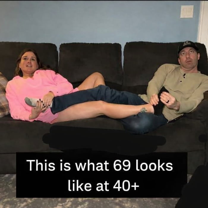 A photograph of a man and a woman sitting on a dark brown couch in a casual home setting. The woman, dressed in a pink top and blue jeans, has her legs stretched across the man's lap, while the man, wearing a beige shirt and a cap, holds one of her feet. Both appear relaxed but with slightly awkward expressions. The text overlay at the bottom reads 'This is what 69 looks like at 40+', humorously subverting the sexual connotation of the term '69' by depicting a mundane, non-sexual interaction between an older couple.