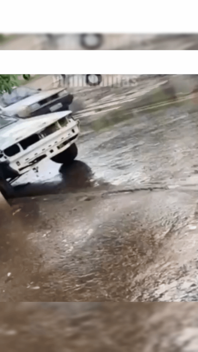 The image depicts a white, possibly old or damaged car partially submerged in murky, muddy water. The car's front end is tilted downward, suggesting it is either stuck or attempting to drive through a flooded or waterlogged outdoor area. In the background, there is some greenery visible, indicating an outdoor setting. The water appears shallow but浑浊, with visible ripples and reflections of the car. The car's front bumper looks worn, adding to the impression of a rugged or challenging situation.