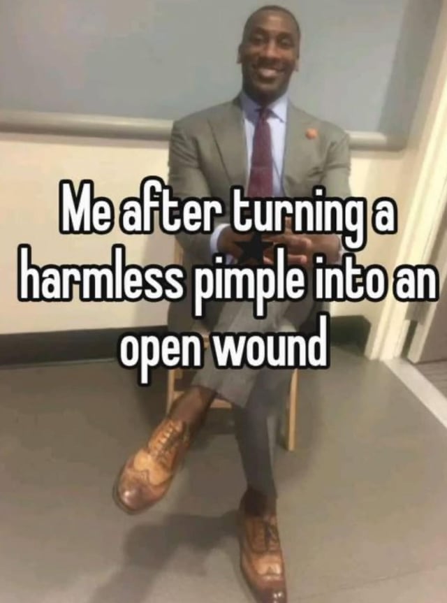 A man in a gray suit, white shirt, maroon tie, and brown leather shoes sits casually on a wooden chair in a room with a gray floor and beige walls. He has a relaxed, confident posture with one leg crossed over the other and hands clasped together. Overlaid text humorously states, "Me after turning a harmless pimple into an open wound," creating a comedic contrast between his self-assured demeanor and the trivial yet relatable skincare mishap.
