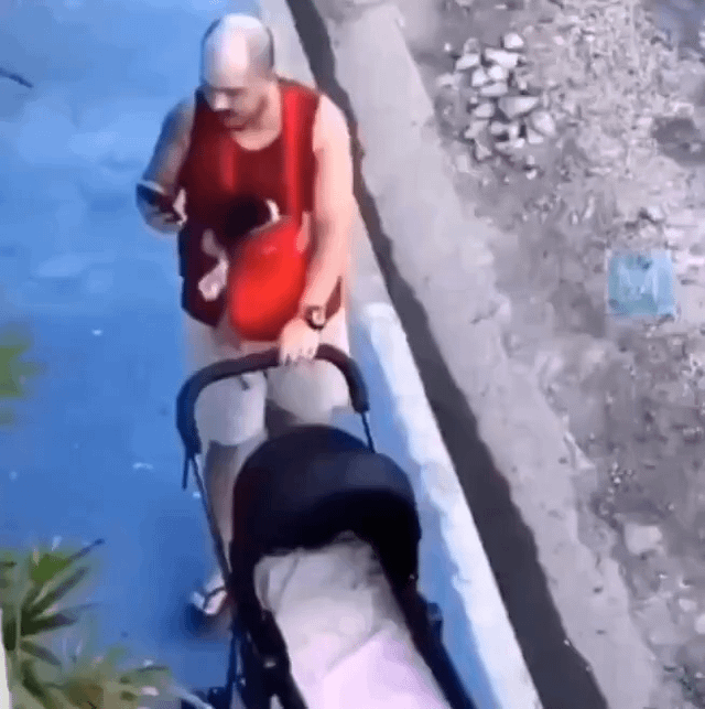 This is a high-angle, first-person perspective photo taken from an elevated position looking down at a sidewalk next to a body of blue water. A bald, mustached man wearing a red sleeveless tank top, white shorts, and flip flops is walking along the sidewalk. He holds a black cat cuddled to his chest like a human baby in one arm, while using his other hand to push a completely empty baby stroller. The humor comes from the absurd, relatable joke of the man treating his pet cat as his child, choosing to carry the cat instead of placing it (or an actual infant) in the stroller, matching the 'me as a parent' framing of the post.