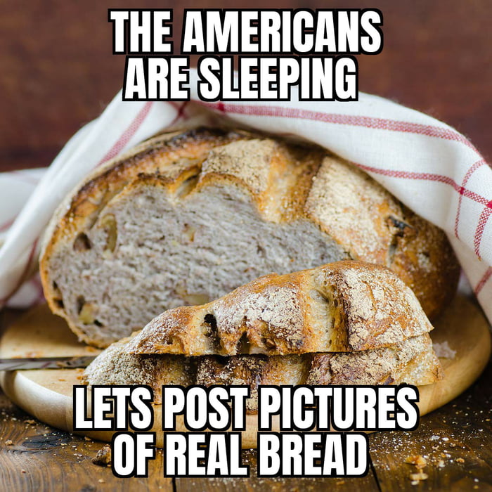The image features a close-up of a rustic, crusty loaf of bread with a textured exterior, partially sliced to reveal its soft, grainy interior. The bread is placed on a wooden board, partially covered by a white cloth with red checkered patterns. Two lines of bold, uppercase text are overlaid: 'THE AMERICANS ARE SLEEPING' at the top and 'LETS POST PICTURES OF REAL BREAD' at the bottom, creating a playful joke that humorously contrasts perceptions of American bread quality with the visually appealing 'real bread' shown.