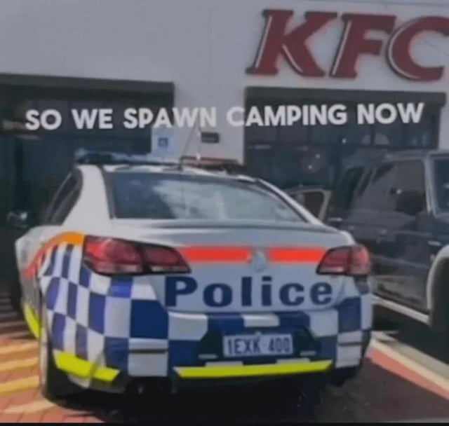 This meme features a photograph of an Australian checkered livery police car parked directly in front of a KFC restaurant outlet. White overlaid text on the upper part of the image reads 'SO WE SPAWN CAMPING NOW'. The joke relies on the video gaming term 'spawn camping', which refers to the practice of waiting at the location where opposing players re-enter the game after being eliminated to easily attack them. It humorously implies that police are waiting at KFC to catch people who commit minor traffic or parking violations while visiting the popular fast food chain, framing the police as using the same cheap tactic as unsportsmanlike gamers.