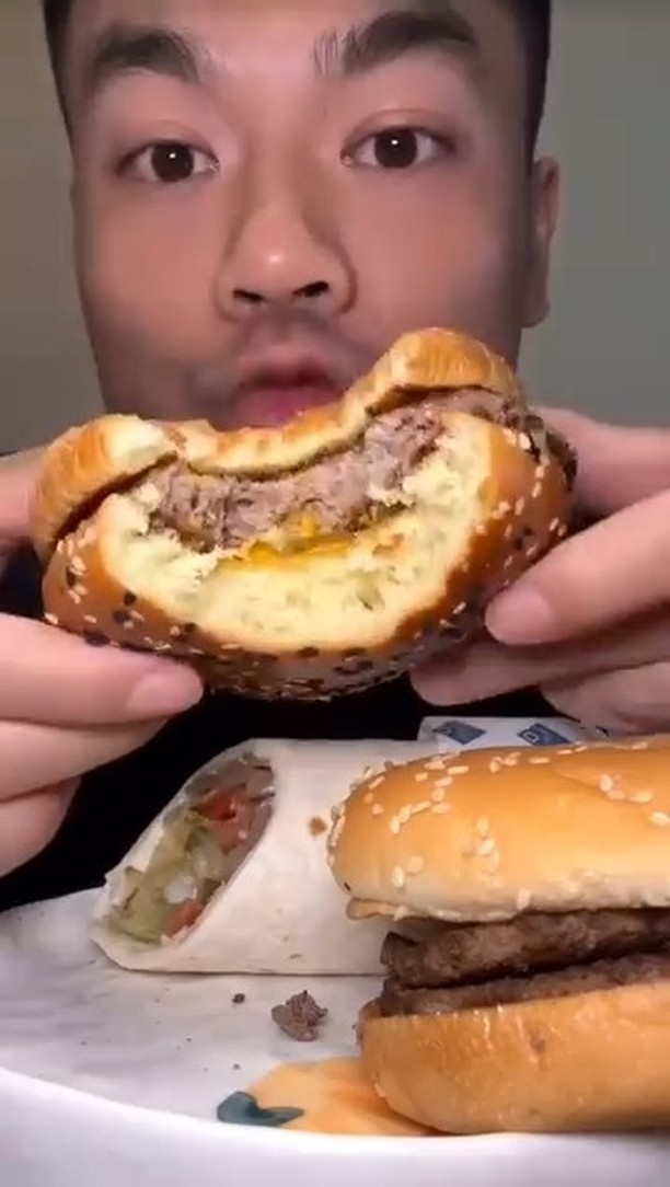 A close-up image of a person holding a partially eaten burger with a sesame seed bun, showcasing the juicy patty and melted cheese inside. In front of them, a white plate holds another intact burger (appearing to have two patties) and a wrapped burrito/wrap with visible vegetables like lettuce and tomatoes. The scene suggests a casual fast food meal, emphasizing the enjoyment of eating.