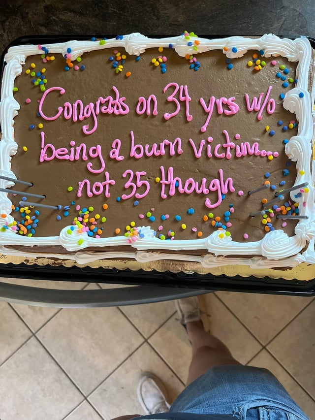 A rectangular chocolate cake with white decorative frosting borders and colorful sprinkles. The cake features pink icing text that reads: 'Congrats on 34 yrs w/o being a burn victim... not 35 though.' A person's legs and feet in white shoes and denim shorts are visible at the bottom, suggesting the photo is taken from a first-person perspective looking down at the cake placed on a tiled floor.