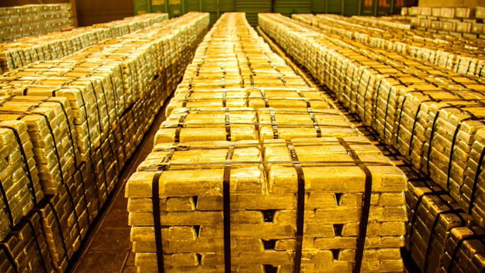 The image depicts a large-scale storage facility filled with neatly stacked gold bars. Numerous rectangular gold bars, secured with black straps, are arranged in parallel rows that extend into the distance, creating a sense of depth and vastness. The gold bars exhibit a metallic sheen under the lighting, highlighting their reflective surface. The background features green industrial containers or walls, contributing to the warehouse-like atmosphere. The overall scene conveys a sense of immense wealth and organized storage of precious metals.
