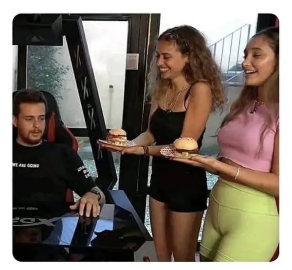A scene depicting a man sitting in a gaming chair at a desk, wearing a black shirt with partial text 'WE ARE GOING' visible. Two women stand in front of him, each holding a burger on a paper plate. The woman on the left is dressed in a black tank top and black shorts, while the woman on the right wears a pink crop top and light green shorts. Both women appear to be smiling as they present the burgers. The setting includes a window with an outdoor view, suggesting an indoor casual environment, possibly a home or gaming room.
