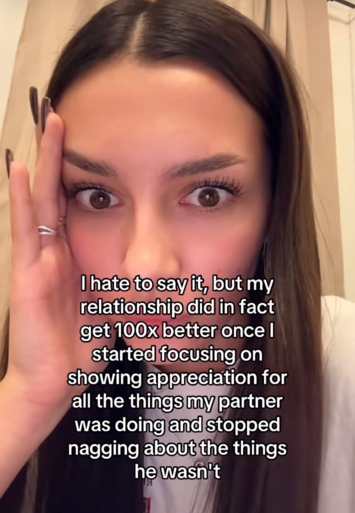 This is a close-up selfie of a young woman with long dark brown hair, dark manicured nails, and silver rings on her fingers, holding her left hand up to the side of her face while looking directly at the camera with a wide-eyed, slightly sheepish, reluctant expression. Overlaid on the lower half of the image is white text with a black outline that reads: 'I hate to say it, but my relationship did in fact get 100x better once I started focusing on showing appreciation for all the things my partner was doing and stopped nagging about the things he wasn\'t.' The accompanying original post title highlights that the woman is clearly reluctant to admit this life and relationship lesson publicly.
