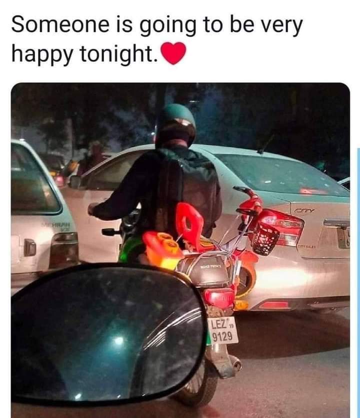 This is a candid nighttime traffic photograph taken from inside another vehicle, with a car side mirror visible in the lower left foreground. A man wearing a teal helmet, seen from the back, rides a motorbike in stopped traffic. A small, brightly colored red and orange children's toy motorcycle is strapped to the rear of his motorbike. Text above the photograph reads "Someone is going to be very happy tonight." paired with a red heart emoji, implying the rider is bringing the toy home as a gift for a child who will be excited to receive it.