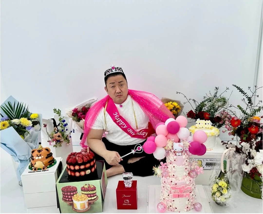 A person sitting cross-legged on the floor, surrounded by festive birthday decorations. They are wearing a white shirt, a pink sash with 'Happy Birthday' written on it, a small tiara, and a pink tulle veil. In their hand, they hold a small magic wand with a heart-shaped top. Surrounding them are various birthday items: a strawberry-topped cake, a tall pink and white cake decorated with Hello Kitty figures and pink/white balloons, a tiger-shaped cake, multiple bouquets of flowers (yellow, red, and mixed colors), and gift boxes. The scene is vibrant and celebratory, indicating a birthday celebration.