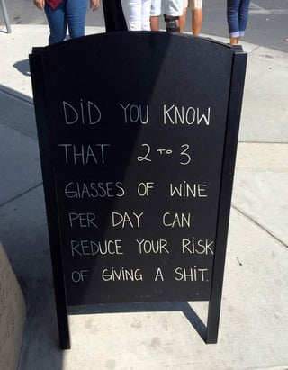 This image shows a black chalkboard sidewalk sandwich board sign placed outdoors on a concrete sidewalk, with the lower legs of several standing people visible in the background. The sign has handwritten white text that creates a humorous, subversive joke: it sets up the expectation of sharing a legitimate health fact about daily wine consumption, but ends with the crude punchline that 2 to 3 glasses of wine a day reduces your risk of "giving a shit", meaning it helps you stop caring about stressful, trivial problems. This aligns with the original Reddit post title about improving mental health through wine. The sign contains a typo, spelling "glasses" as "giasses".